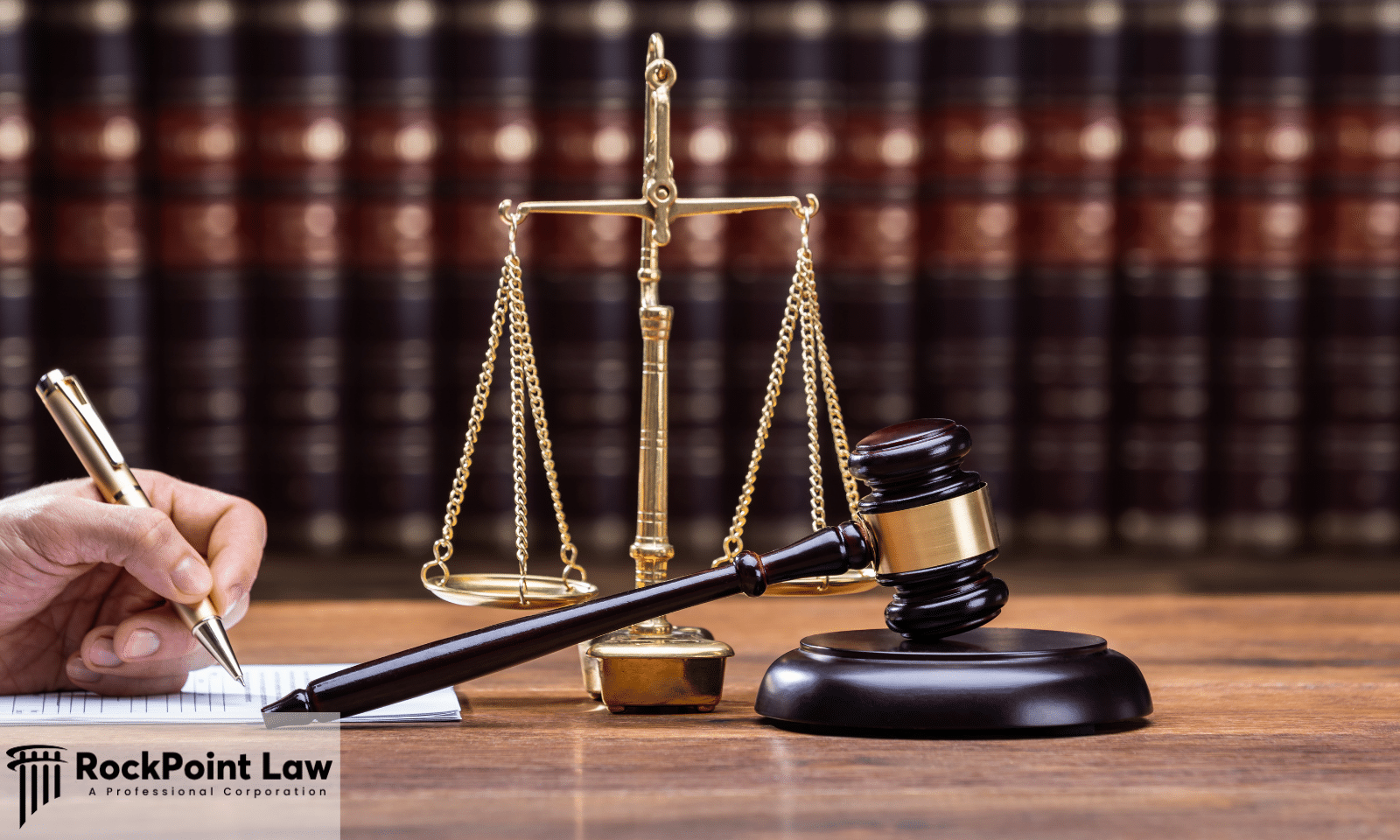 Wooden judge's gavel and brass scales of justice on a desk beside a person signing legal documents, with law books in the background.