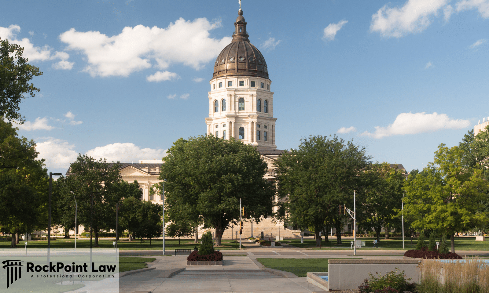 Kansas State Capitol building in Topeka surrounded by trees and landscaped grounds under a blue sky.