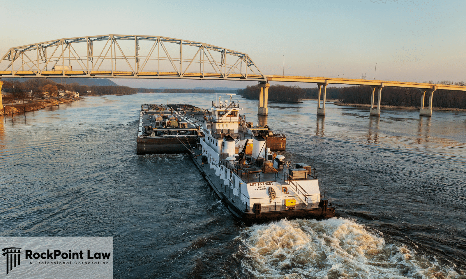 Commercial towboat pushing barges along the Mississippi River beneath a steel bridge during sunset.