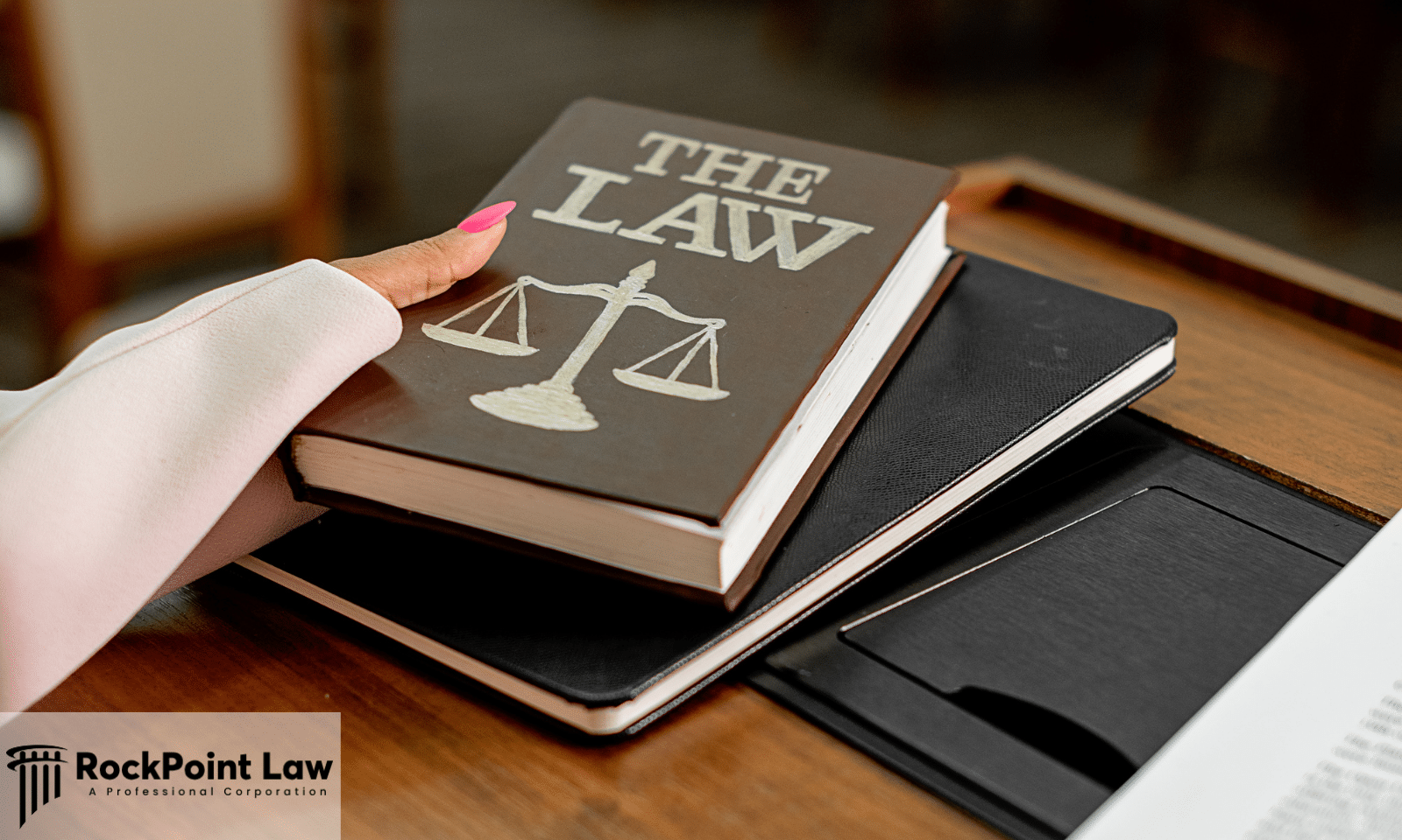 Person holding a law book labeled "The Law" with scales of justice on the cover beside legal documents.