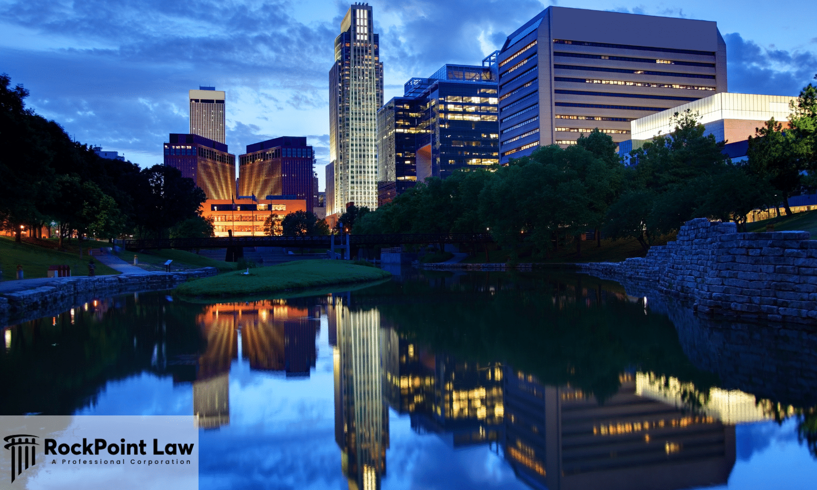 Omaha, Nebraska skyline reflected in a calm river at dusk with illuminated downtown buildings.