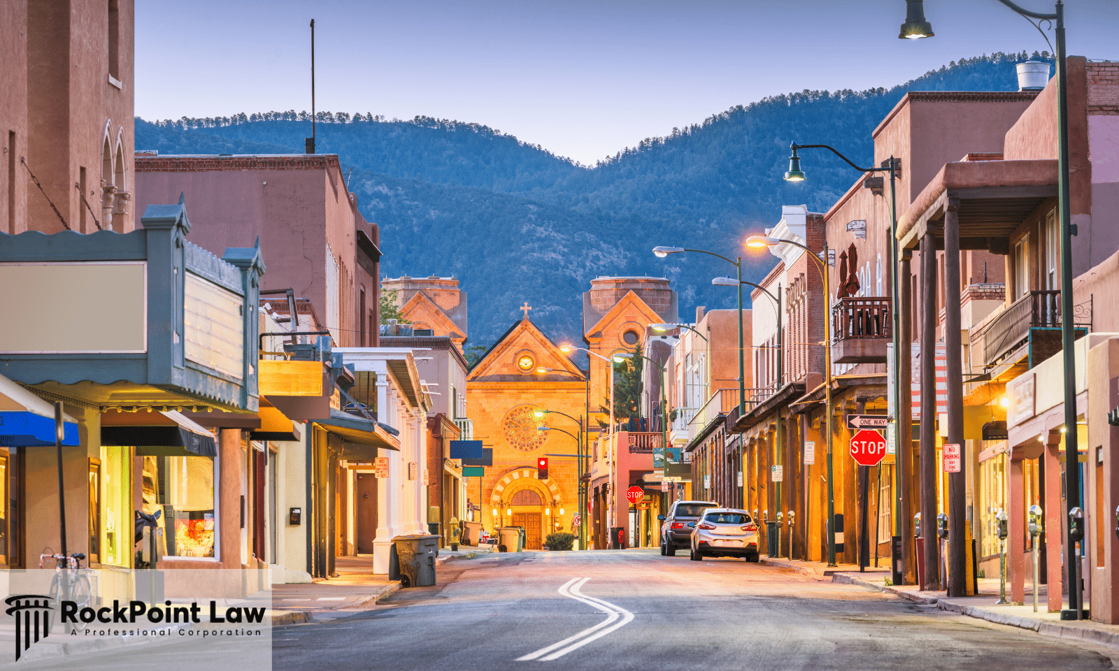 Historic downtown Santa Fe street at dusk with adobe storefronts, the cathedral, and mountains beyond.
